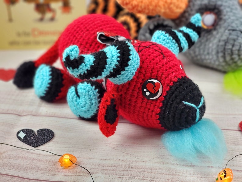 Peut inclure: Une cr&eacute;ature rouge faite &agrave; la main au crochet avec des accents noirs et bleus. Le jouet en peluche a un museau noir, une touffe bleue et un &oelig;il bouton avec un c&oelig;ur bris&eacute;. D'autres jouets au crochet sont en arri&egrave;re-plan.