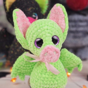 May include: A green crocheted bat with pink ears and a pink nose. The bat has large purple eyes and a pink ribbon around its neck.