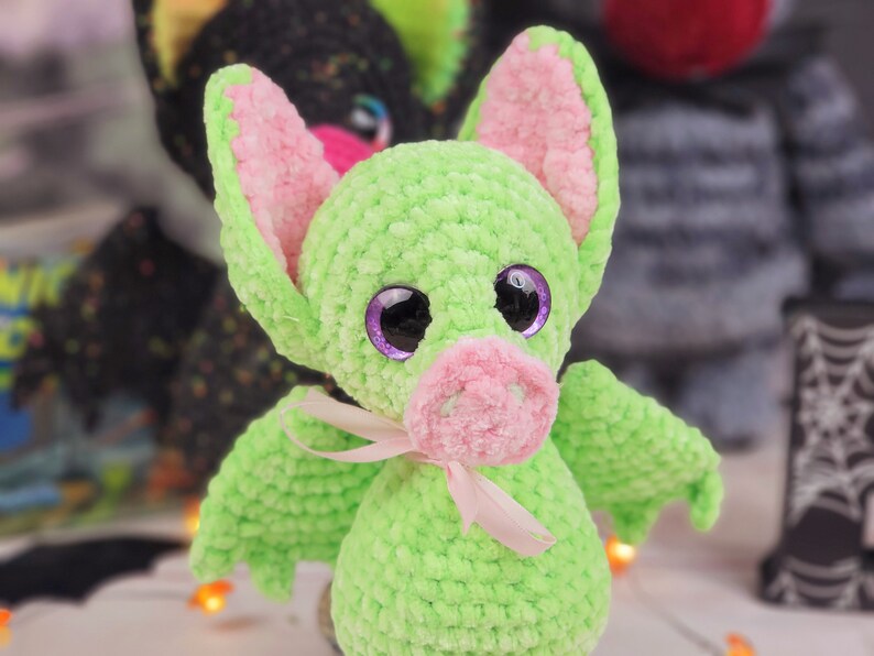 May include: A green crocheted bat with pink ears and a pink nose. The bat has large purple eyes and a pink ribbon around its neck.