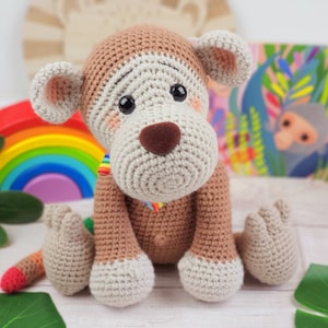 May include: A crocheted monkey toy with brown and white fur, sitting on a white surface. The monkey has a brown nose and large, blue eyes.