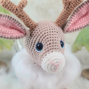 May include: A crocheted reindeer head with brown antlers, large blue eyes, and pink-lined ears. The reindeer has a white muzzle and a fluffy white collar. The background is blurred, suggesting a winter or holiday theme.