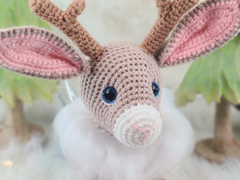 May include: A crocheted reindeer head with brown antlers, large blue eyes, and pink-lined ears. The reindeer has a white muzzle and a fluffy white collar. The background is blurred, suggesting a winter or holiday theme.