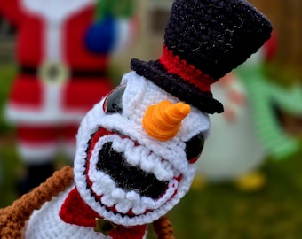 May include: A crocheted snowman with a black top hat, orange carrot nose, and a wide, toothy grin. The snowman is wearing a red scarf and has brown yarn arms.