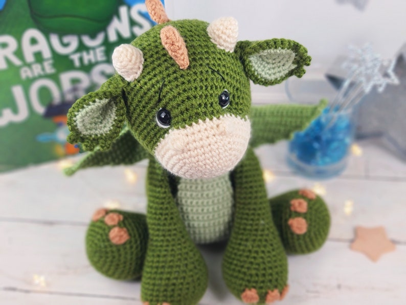 May include: A green crocheted dragon toy with a cream-coloured snout and brown spots on its paws. The dragon has a small, orange horn on its head and large, black eyes.