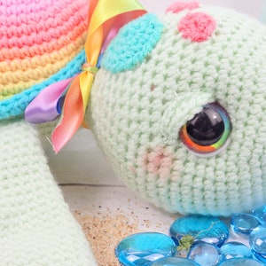 Puede incluir: Un juguete de tortuga de crochet con una concha de arco&iacute;ris y un cuerpo verde. La tortuga tiene un ojo grande y colorido y est&aacute; tumbada sobre una superficie blanca con piedras de vidrio azules.
