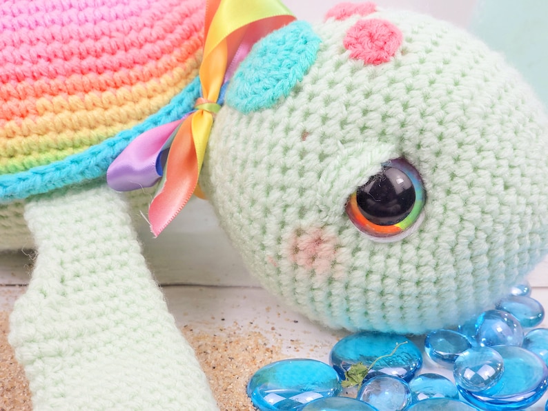Puede incluir: Un juguete de tortuga de crochet con una concha de arco&iacute;ris y un cuerpo verde. La tortuga tiene un ojo grande y colorido y est&aacute; tumbada sobre una superficie blanca con piedras de vidrio azules.