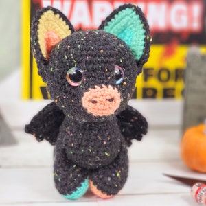 May include: A black crocheted bat with colorful ears, pink nose, and sparkly eyes. The bat is standing on a white surface with a pumpkin and a warning sign in the background.