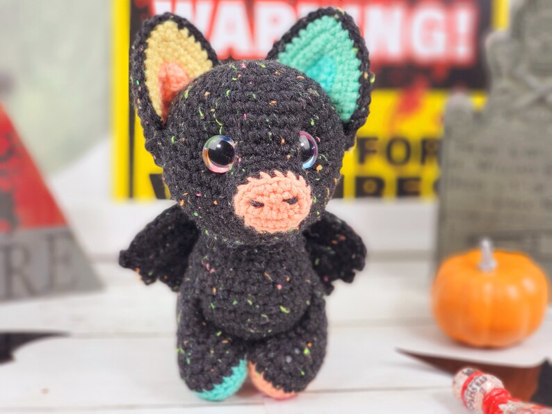May include: A black crocheted bat with colorful ears, pink nose, and sparkly eyes. The bat is standing on a white surface with a pumpkin and a warning sign in the background.