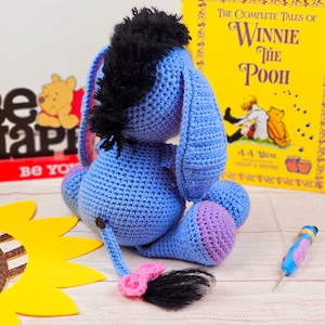 May include: A blue crocheted Eeyore plush toy with a black mane and a pink tail. The toy is sitting on a wooden surface with a sunflower and a book titled "The Complete Tales of Winnie the Pooh" in the background.
