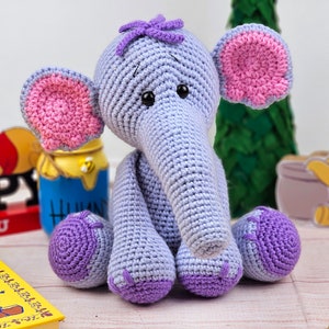 May include: A crocheted purple and pink elephant toy with a long trunk and big ears. The elephant is sitting on a wooden surface.
