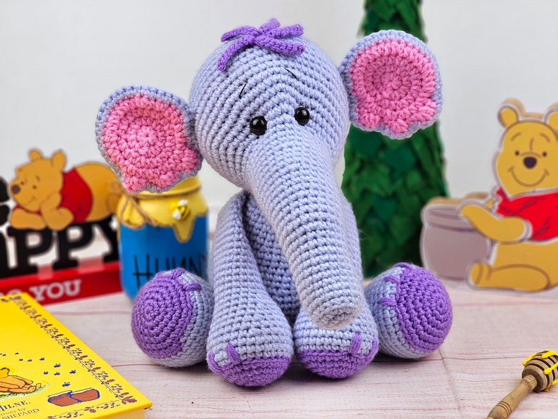 May include: A crocheted purple and pink elephant toy with a long trunk and big ears. The elephant is sitting on a wooden surface.