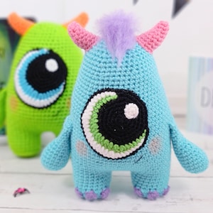Può includere: Due mostri di peluche all'uncinetto con occhi grandi. Un mostro è verde con corna arancioni e l'altro è blu con corna rosa e pelliccia viola sulla testa.
