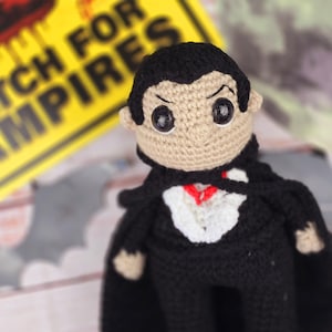 May include: A crocheted vampire doll wearing a black cape and a white shirt with a red tie. The doll has black hair, brown eyes, and a stitched-on smile. The doll is standing in front of a yellow sign that reads "Watch for Vampires".