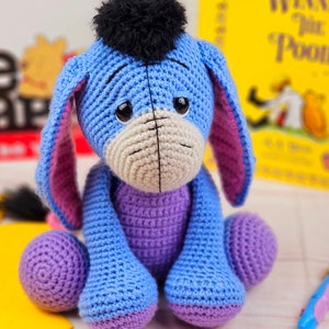May include: A crocheted stuffed toy donkey with blue and purple fur, black yarn hair, and large brown eyes. The donkey is sitting on a white surface.