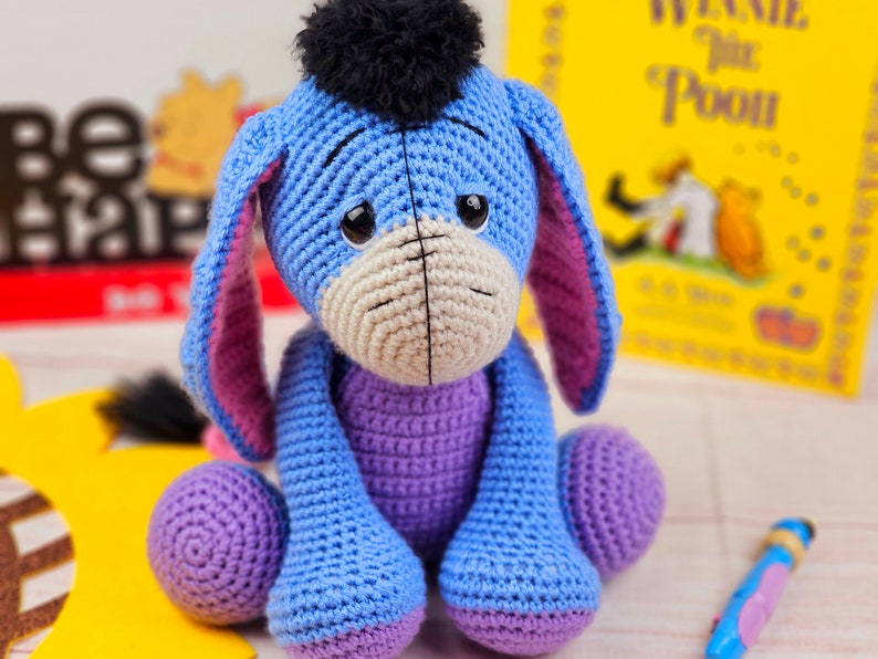 May include: A crocheted stuffed toy donkey with blue and purple fur, black yarn hair, and large brown eyes. The donkey is sitting on a white surface.