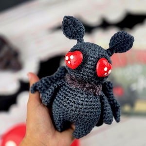 May include: A handmade crochet moth with dark gray yarn. The moth has large, heart-shaped red eyes with white dots, small wings, and two rounded antennae. The moth is a Halloween or gothic-themed item.