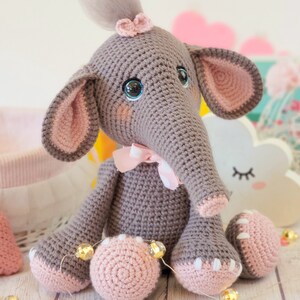 May include: A crocheted gray elephant toy with pink ears, a pink bow, and blue eyes. The elephant is sitting on a white surface with pink hearts and gold string lights.