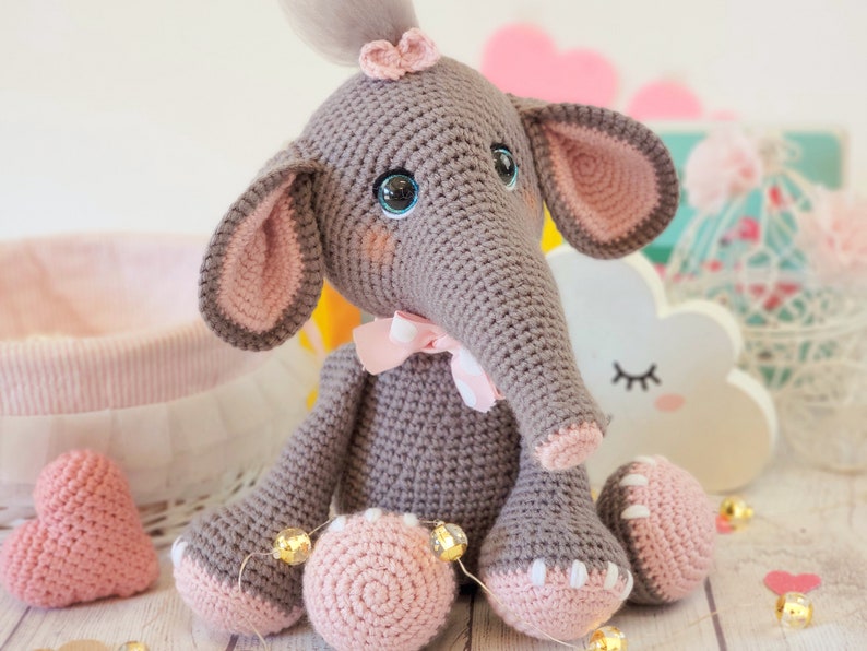May include: A crocheted gray elephant toy with pink ears, a pink bow, and blue eyes. The elephant is sitting on a white surface with pink hearts and gold string lights.