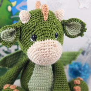 May include: A green crocheted dragon toy with a light brown nose, orange horns, and light green ears. The dragon has black eyes and is sitting on a white surface.