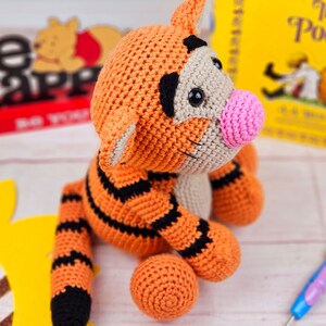 May include: A crocheted orange and black tiger plush toy with a pink nose. The toy is sitting on a white surface with a sunflower and a book titled "The Pooh" in the background.