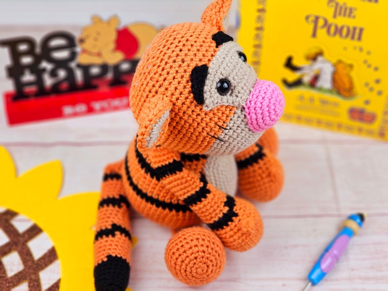 May include: A crocheted orange and black tiger plush toy with a pink nose. The toy is sitting on a white surface with a sunflower and a book titled "The Pooh" in the background.
