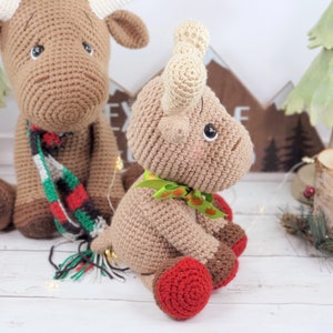 May include: Two crocheted moose toys, one wearing a red and green scarf and the other with a green bow. Both are sitting on a white wooden surface.