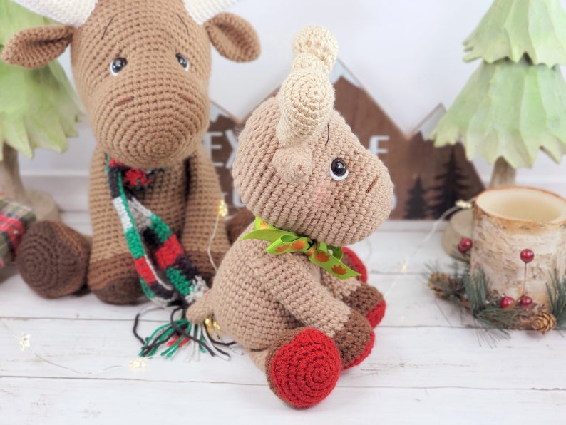 May include: Two crocheted moose toys, one wearing a red and green scarf and the other with a green bow. Both are sitting on a white wooden surface.