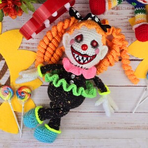 May include: A crocheted clown doll with orange hair, a black and green dress, and a white face with a red nose and a wide, toothy grin. The doll is lying on a wooden surface with a sunflower and lollipops in the background.