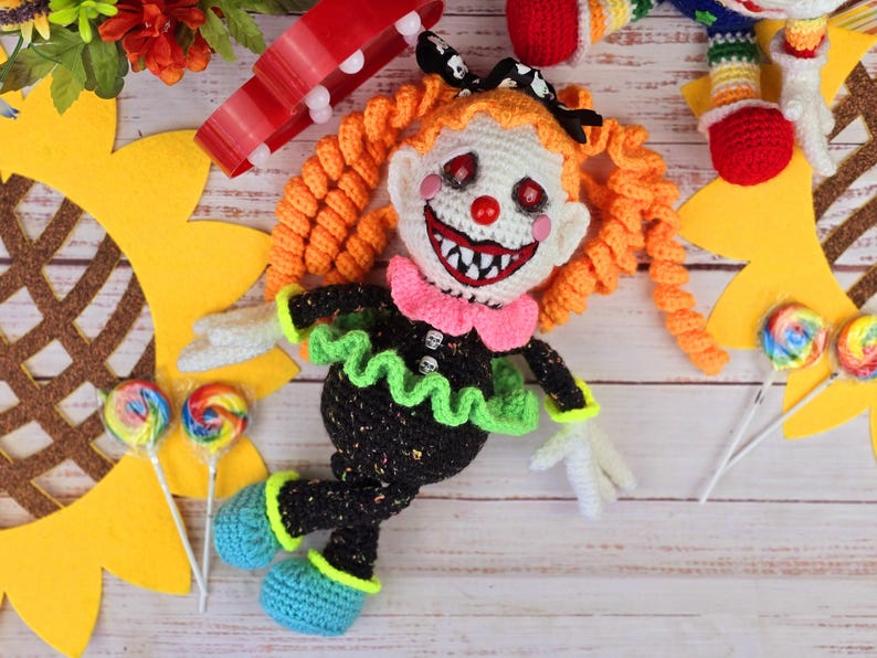 May include: A crocheted clown doll with orange hair, a black and green dress, and a white face with a red nose and a wide, toothy grin. The doll is lying on a wooden surface with a sunflower and lollipops in the background.