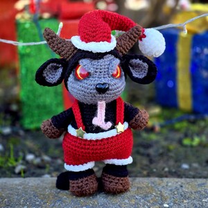 Puede incluir: Una cabra de crochet gris y negro que lleva un gorro de Pap&aacute; Noel rojo con ribete blanco y un traje de Pap&aacute; Noel rojo y blanco con tirantes negros y estrellas doradas.
