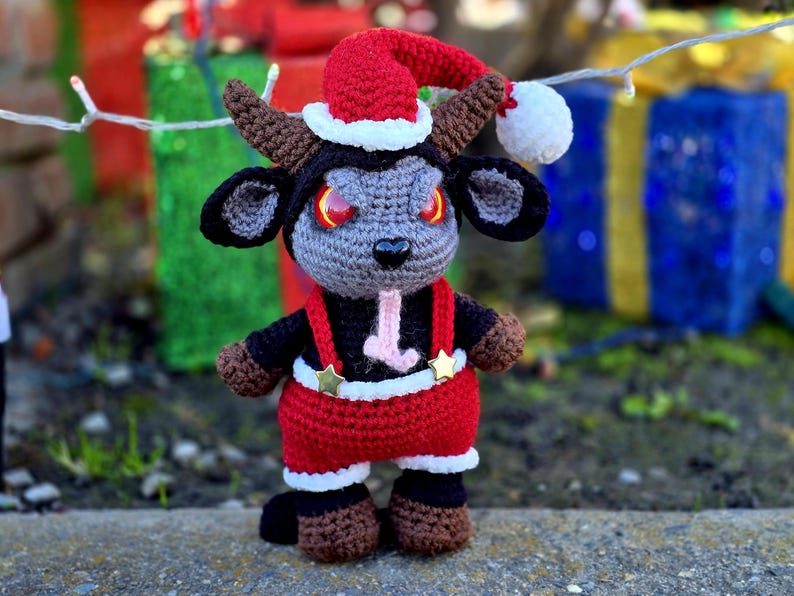 Puede incluir: Una cabra de crochet gris y negro que lleva un gorro de Pap&aacute; Noel rojo con ribete blanco y un traje de Pap&aacute; Noel rojo y blanco con tirantes negros y estrellas doradas.