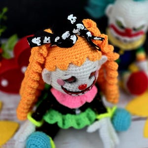 May include: A crocheted clown doll with orange hair, a black bow with white ghost designs, and a white face with a red nose and pink lips. The doll is wearing a black and green outfit with a pink collar.