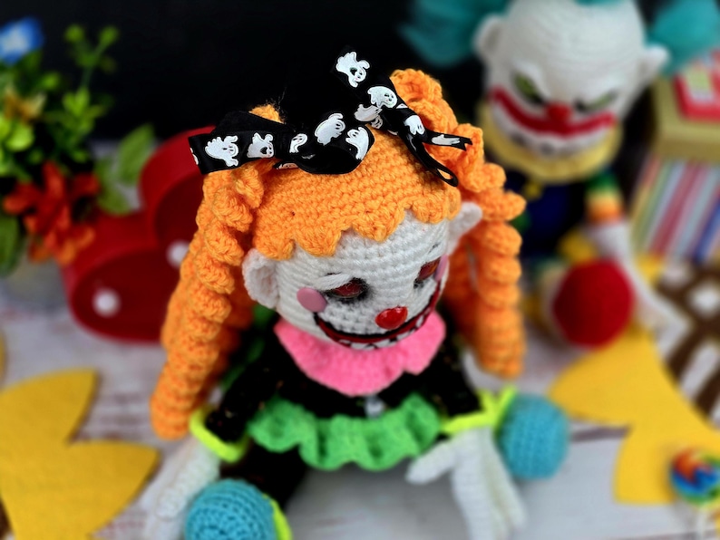 May include: A crocheted clown doll with orange hair, a black bow with white ghost designs, and a white face with a red nose and pink lips. The doll is wearing a black and green outfit with a pink collar.