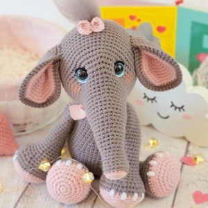May include: A gray crocheted elephant toy with pink ears, a pink bow on its head, and blue eyes. The elephant is sitting on a white wooden surface with pink hearts and a white cloud with a smiley face.