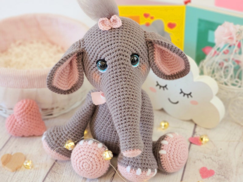 May include: A gray crocheted elephant toy with pink ears, a pink bow on its head, and blue eyes. The elephant is sitting on a white wooden surface with pink hearts and a white cloud with a smiley face.