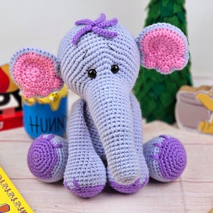 May include: A crocheted purple elephant toy with pink ears and a purple flower on its head. The elephant is sitting on a wooden surface.