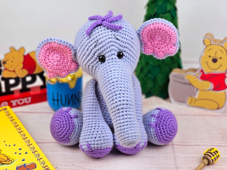 May include: A crocheted purple elephant toy with pink ears and a purple flower on its head. The elephant is sitting on a wooden surface.