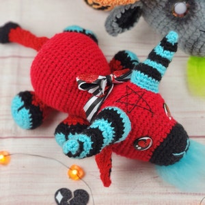 Peut inclure: Une licorne rouge faite &agrave; la main au crochet avec des accents noirs et bleus, un n&oelig;ud ray&eacute; noir et blanc et une queue bleue. La licorne a une corne noire avec des rayures bleues et un pentagramme sur la t&ecirc;te. D'autres cr&eacute;atures au crochet sont en arri&egrave;re-plan.