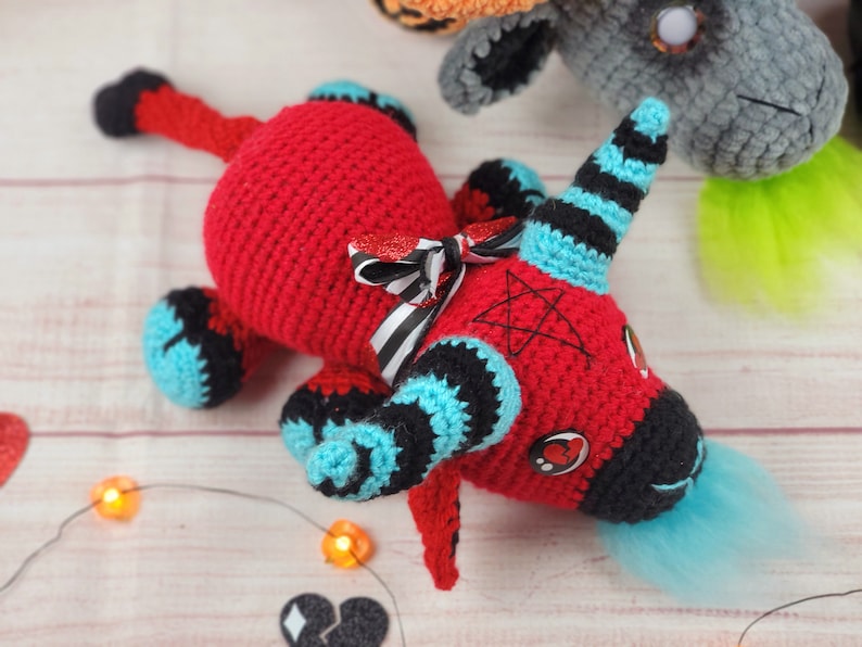 Peut inclure: Une licorne rouge faite &agrave; la main au crochet avec des accents noirs et bleus, un n&oelig;ud ray&eacute; noir et blanc et une queue bleue. La licorne a une corne noire avec des rayures bleues et un pentagramme sur la t&ecirc;te. D'autres cr&eacute;atures au crochet sont en arri&egrave;re-plan.