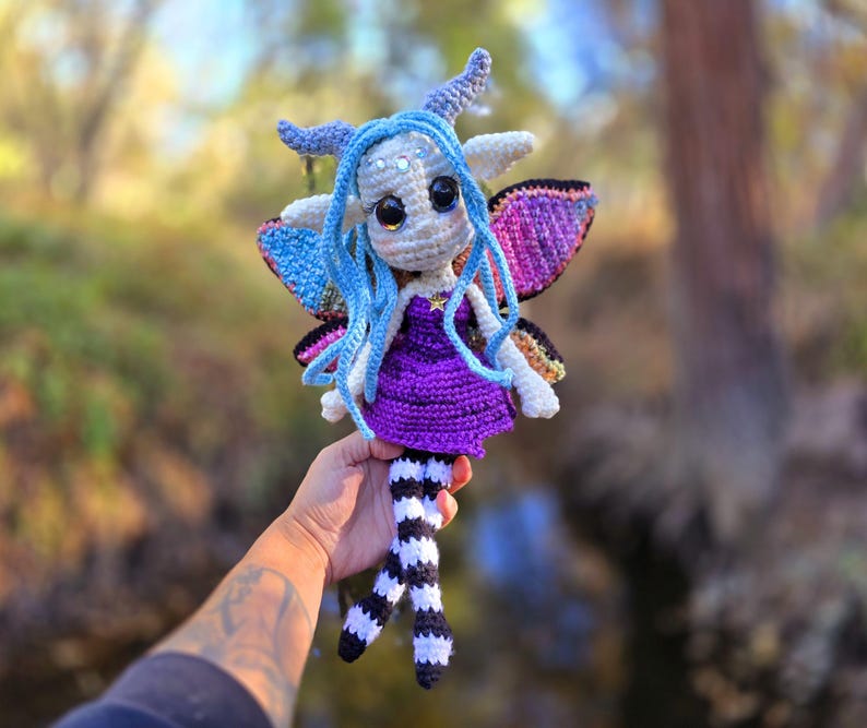 May include: A crocheted fairy doll with blue hair, purple dress, and black and white striped legs. The fairy has iridescent wings and a star on its chest.