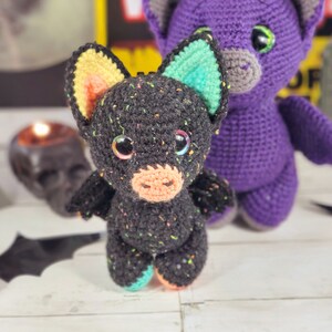 May include: A black crocheted bat with colorful accents, standing next to a purple crocheted bat. Both bats have large eyes and are made with a textured yarn.