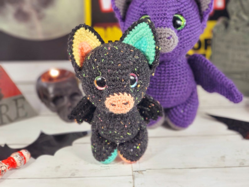 May include: A black crocheted bat with colorful accents, standing next to a purple crocheted bat. Both bats have large eyes and are made with a textured yarn.