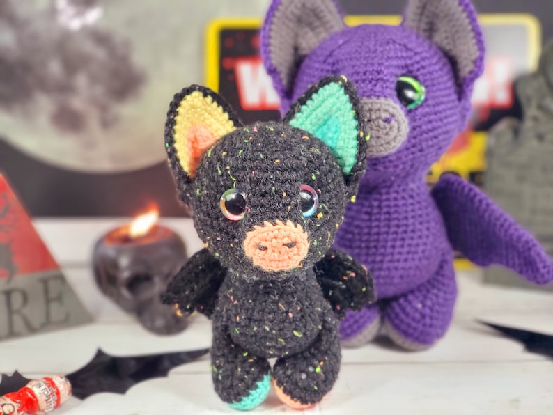 May include: Two crocheted bat toys. One is black with colorful accents and the other is purple. Both have large eyes and wings.