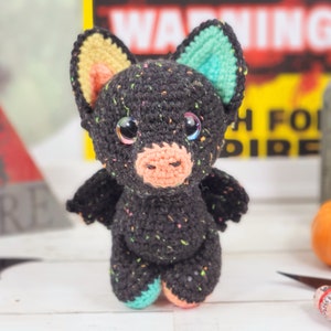 May include: A black crocheted bat with colorful accents. The bat has large eyes, a pink nose, and a speckled black body. The bat is standing on a white surface.