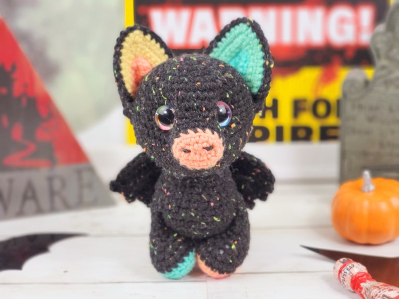 May include: A black crocheted bat with colorful accents. The bat has large eyes, a pink nose, and a speckled black body. The bat is standing on a white surface.
