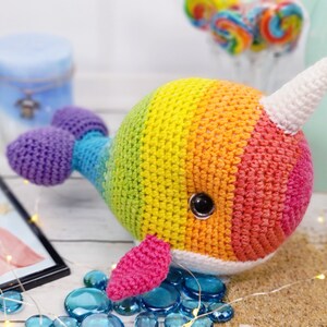 K&ouml;nnte beinhalten: Ein regenbogenfarbenes, geh&auml;keltes Narwal-Pl&uuml;schtier mit einem wei&szlig;en Horn und einer rosa Flosse. Das Spielzeug liegt auf einer wei&szlig;en Oberfl&auml;che mit blauen und wei&szlig;en Dekosteinen.