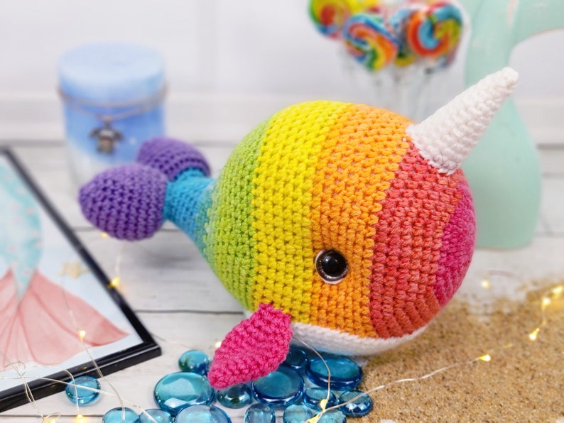 K&ouml;nnte beinhalten: Ein regenbogenfarbenes, geh&auml;keltes Narwal-Pl&uuml;schtier mit einem wei&szlig;en Horn und einer rosa Flosse. Das Spielzeug liegt auf einer wei&szlig;en Oberfl&auml;che mit blauen und wei&szlig;en Dekosteinen.