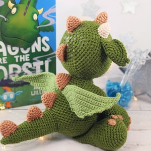 May include: A green crocheted dragon toy with brown and white accents. The dragon has wings and a tail. The toy is sitting on a white surface. The book in the background says "Dragons are the worst!"