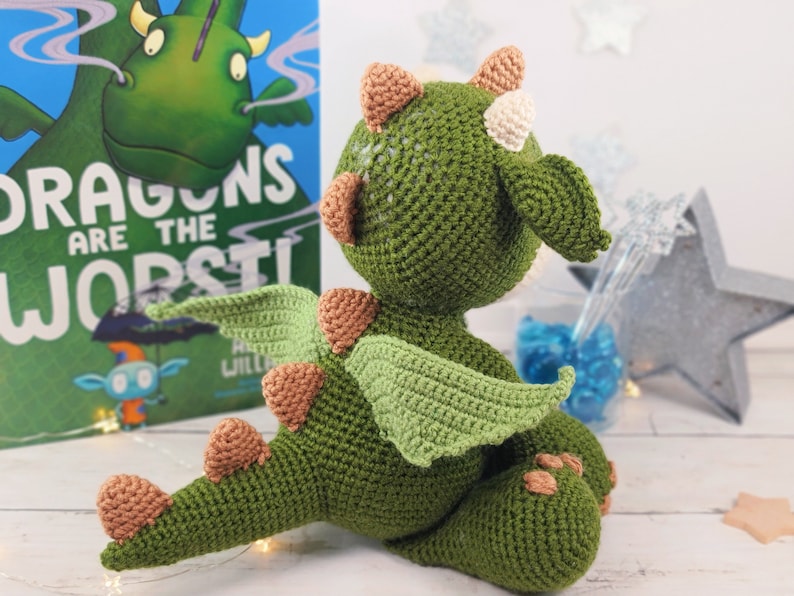 May include: A green crocheted dragon toy with brown and white accents. The dragon has wings and a tail. The toy is sitting on a white surface. The book in the background says "Dragons are the worst!"
