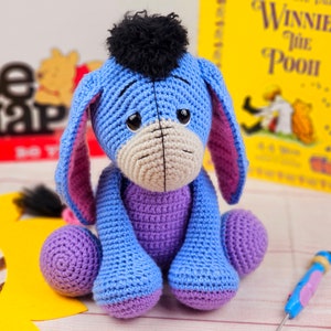 May include: A crocheted blue and purple Eeyore plush toy with a black yarn tail and a black yarn pom-pom on its head. The toy is sitting on a wooden surface with a yellow sunflower and a book titled "Winnie the Pooh" in the background.